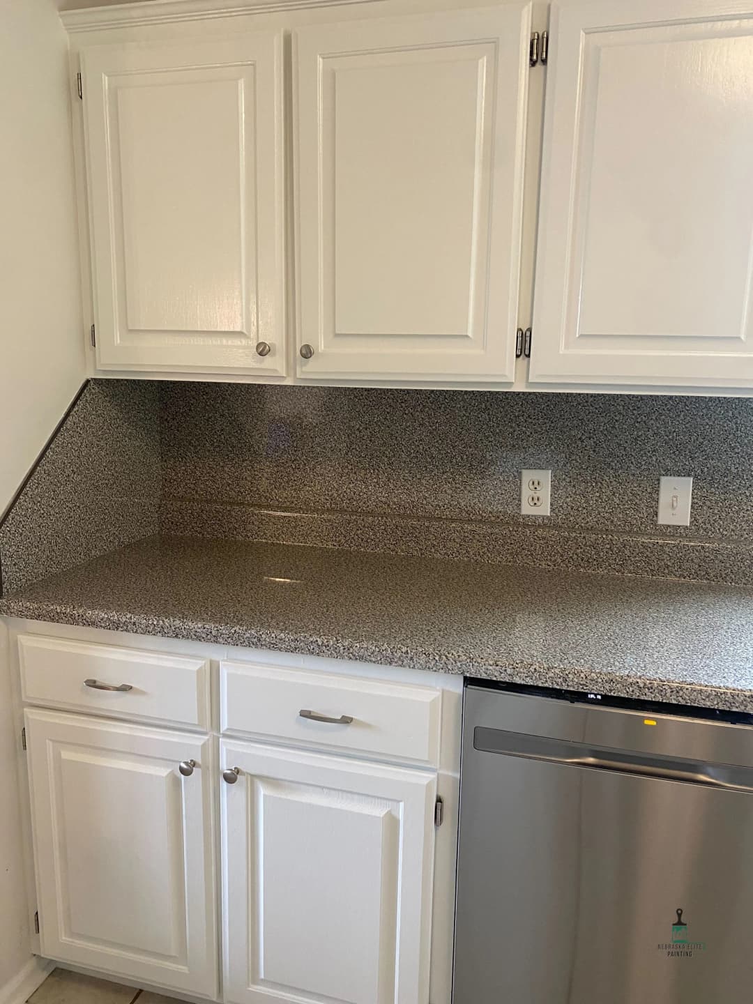 Modern kitchen with white cabinetry and speckled countertops. Stainless steel dishwasher visible.