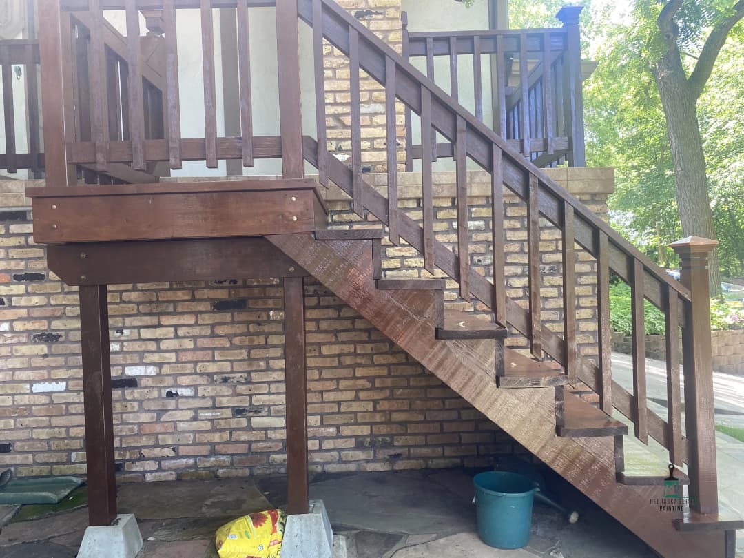 Wooden staircase with railing, leading to a brick house, surrounded by greenery.