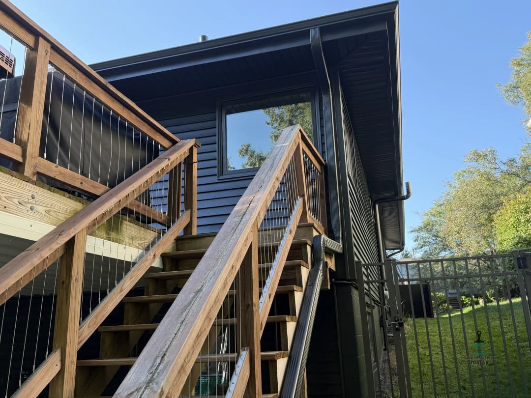 Wooden stairs lead to a modern black house with large windows and green surroundings.