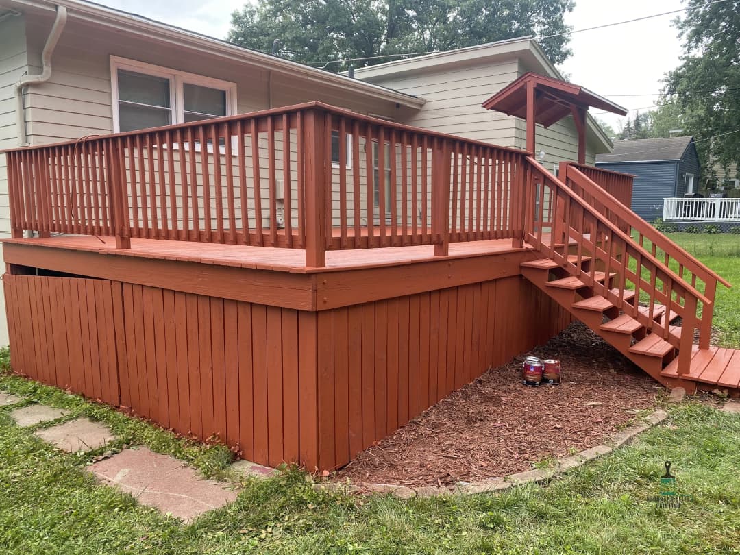 Red wooden deck with railing and stairs, surrounded by green grass and mulch landscaping.