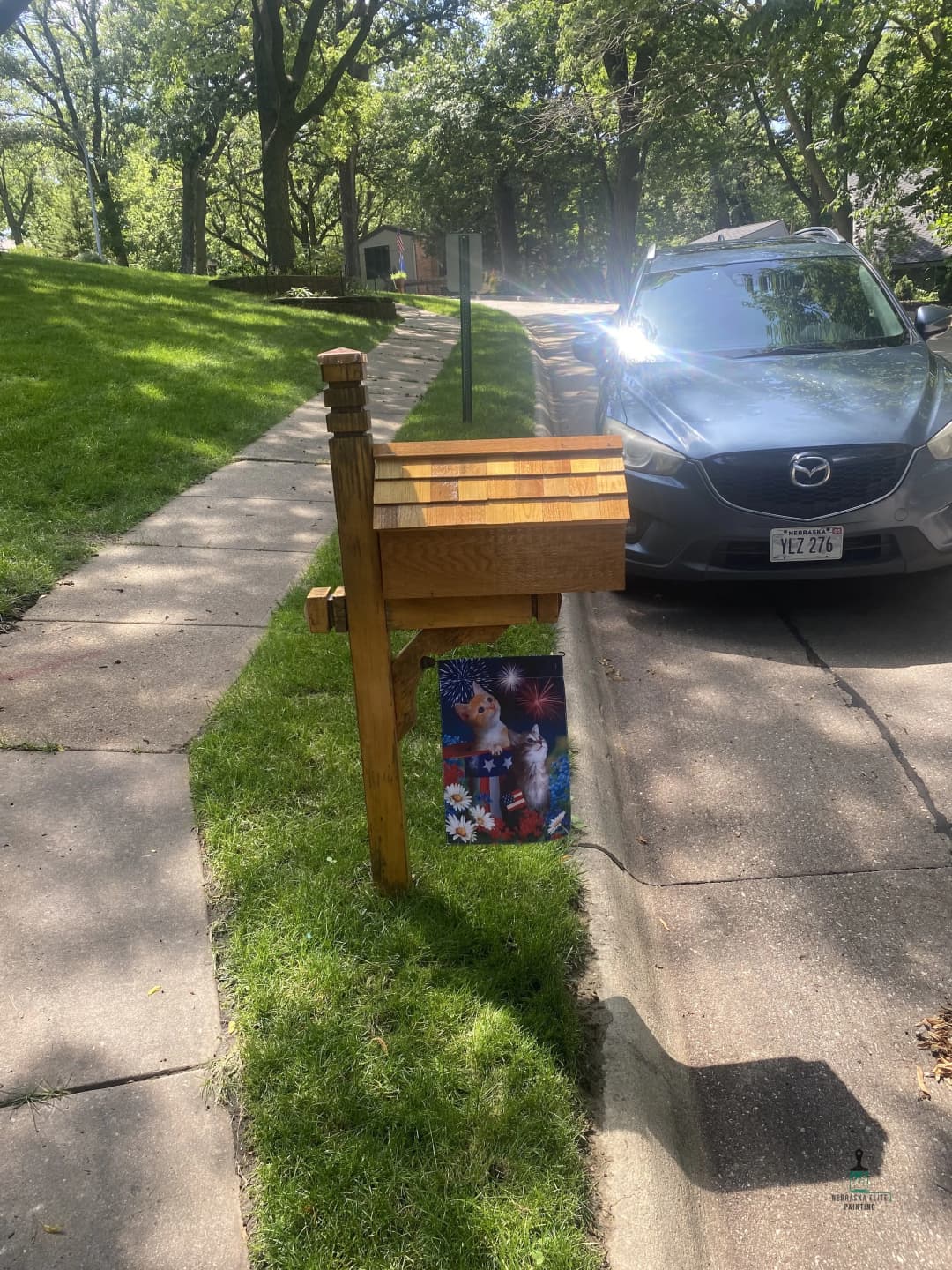 Wooden mailbox with decorative holiday flag beside a car on a sunny residential street.