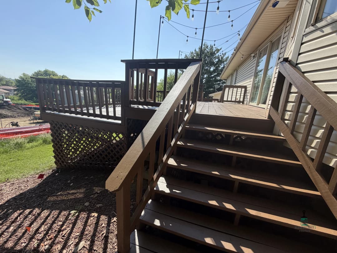 Wooden deck with stairs, surrounded by greenery and decorative string lights, sunny day.