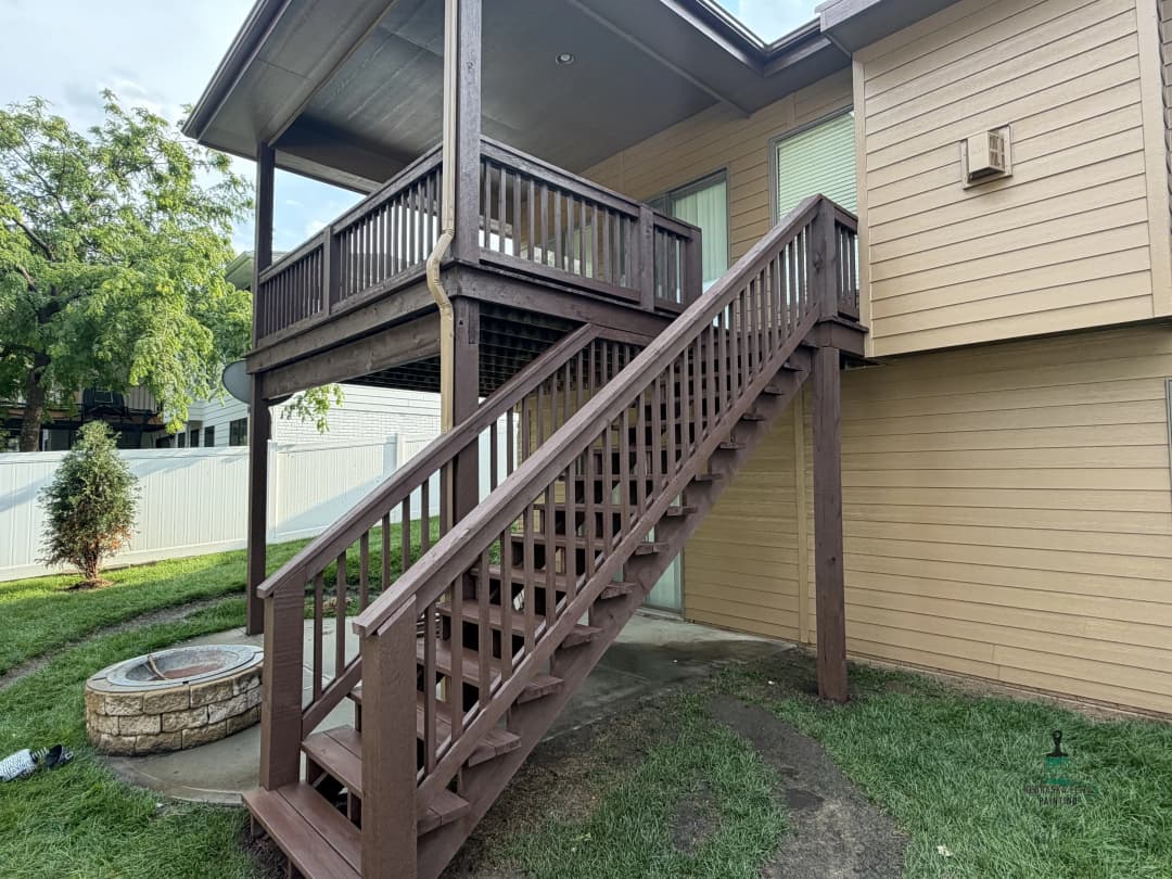 Exterior view of a two-story home with a wooden staircase and balcony, surrounded by greenery.