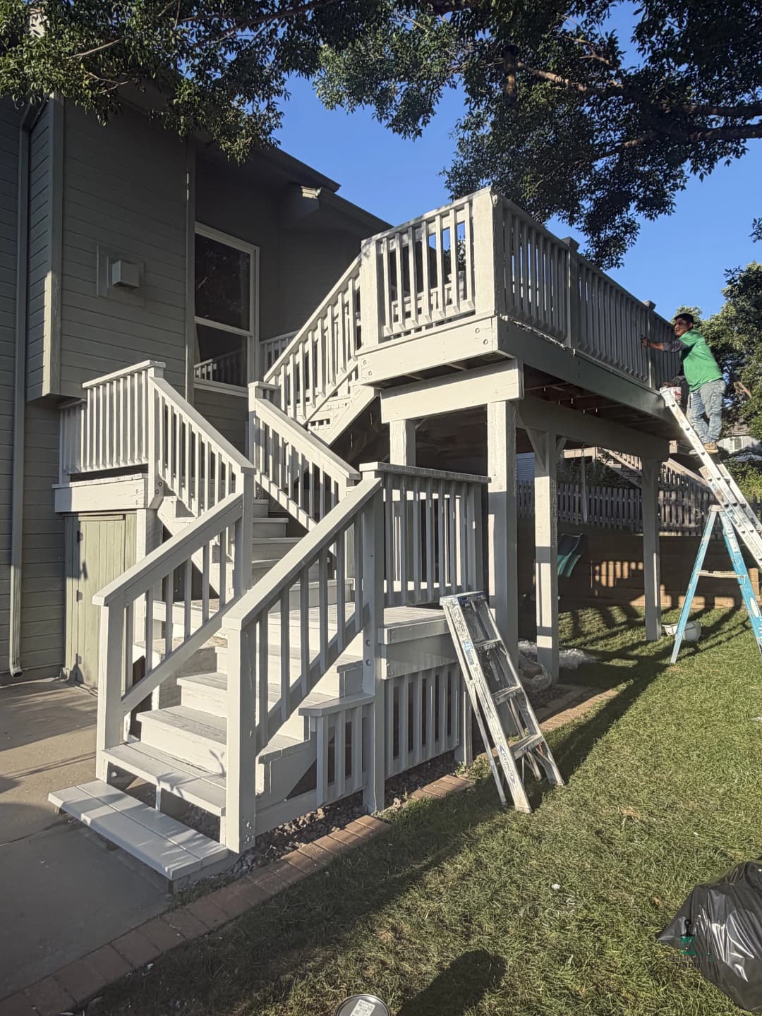 Renovated two-level wooden deck with stairs and workers, surrounded by greenery.