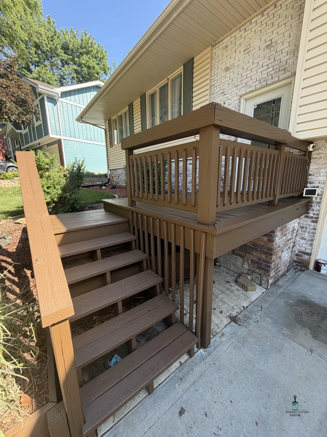 Brown wooden deck with stairs leading to a brick house, surrounded by greenery.