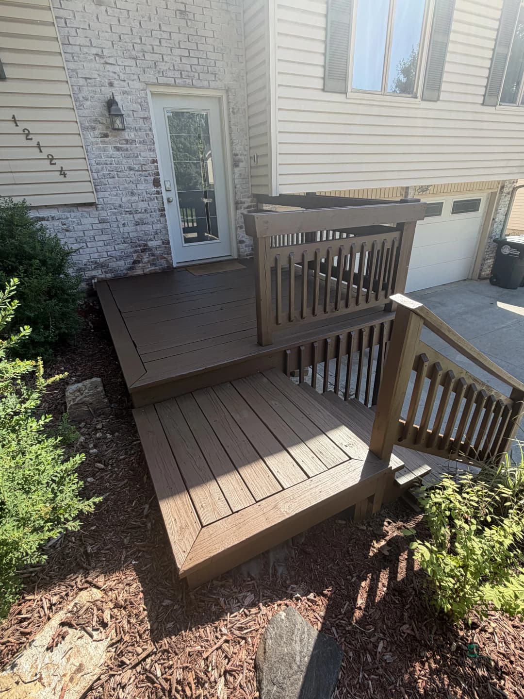 Brown wooden deck with railing, leading to a home entrance, surrounded by shrubs and gravel.
