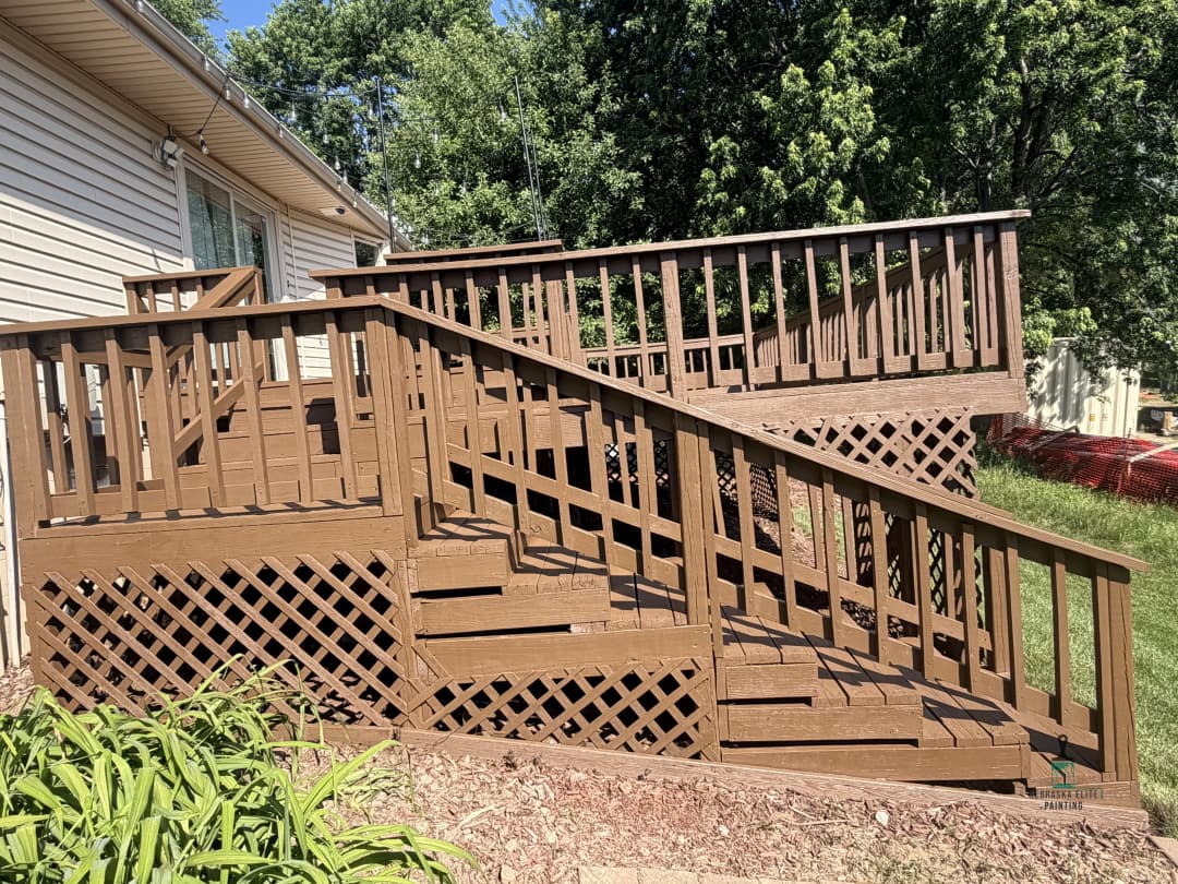 Brown wooden deck with railings and stairs, surrounded by greenery and landscaping.