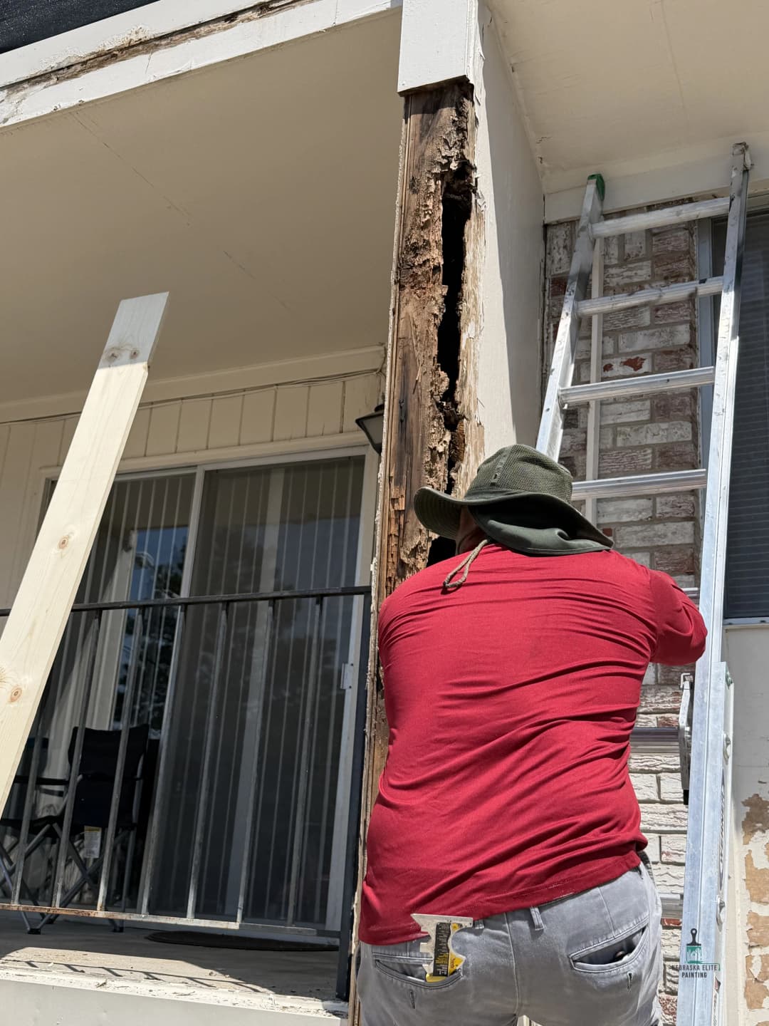 Man repairing damaged wood on a building with a ladder and tools.