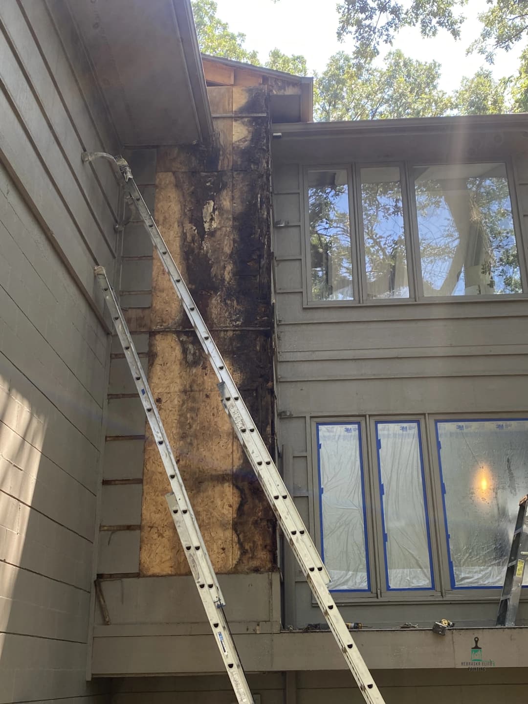 Damaged exterior wall with mold, ladders for repair, and windows covered in plastic.