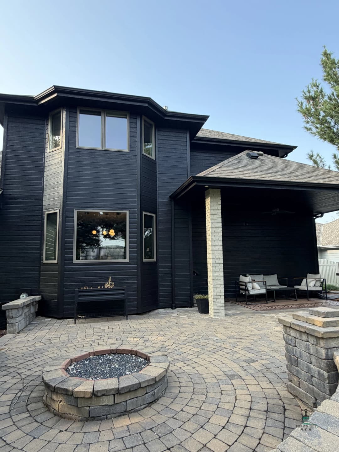 Modern black house with outdoor seating and stone fire pit, surrounded by paver patio.