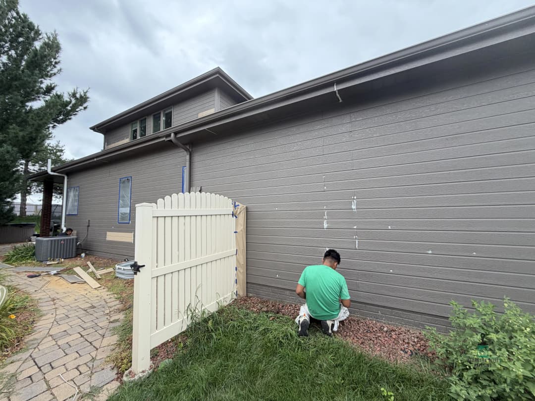 Person kneeling by a gray house wall, tending to landscaping near a white fence.