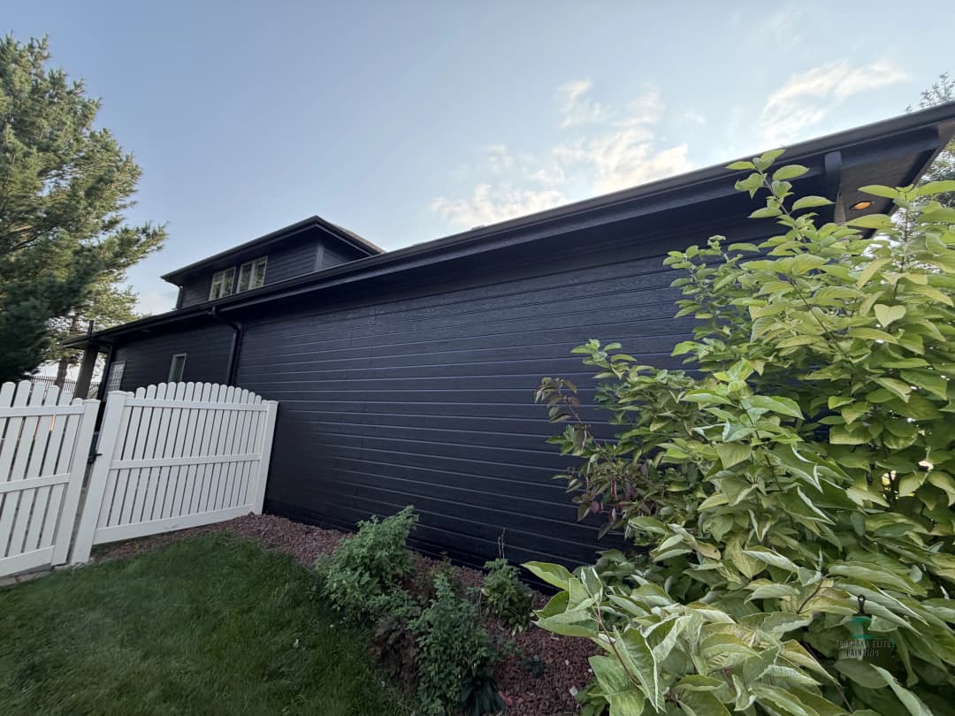 Black wood-paneled house exterior with white picket fence and greenery in the foreground.