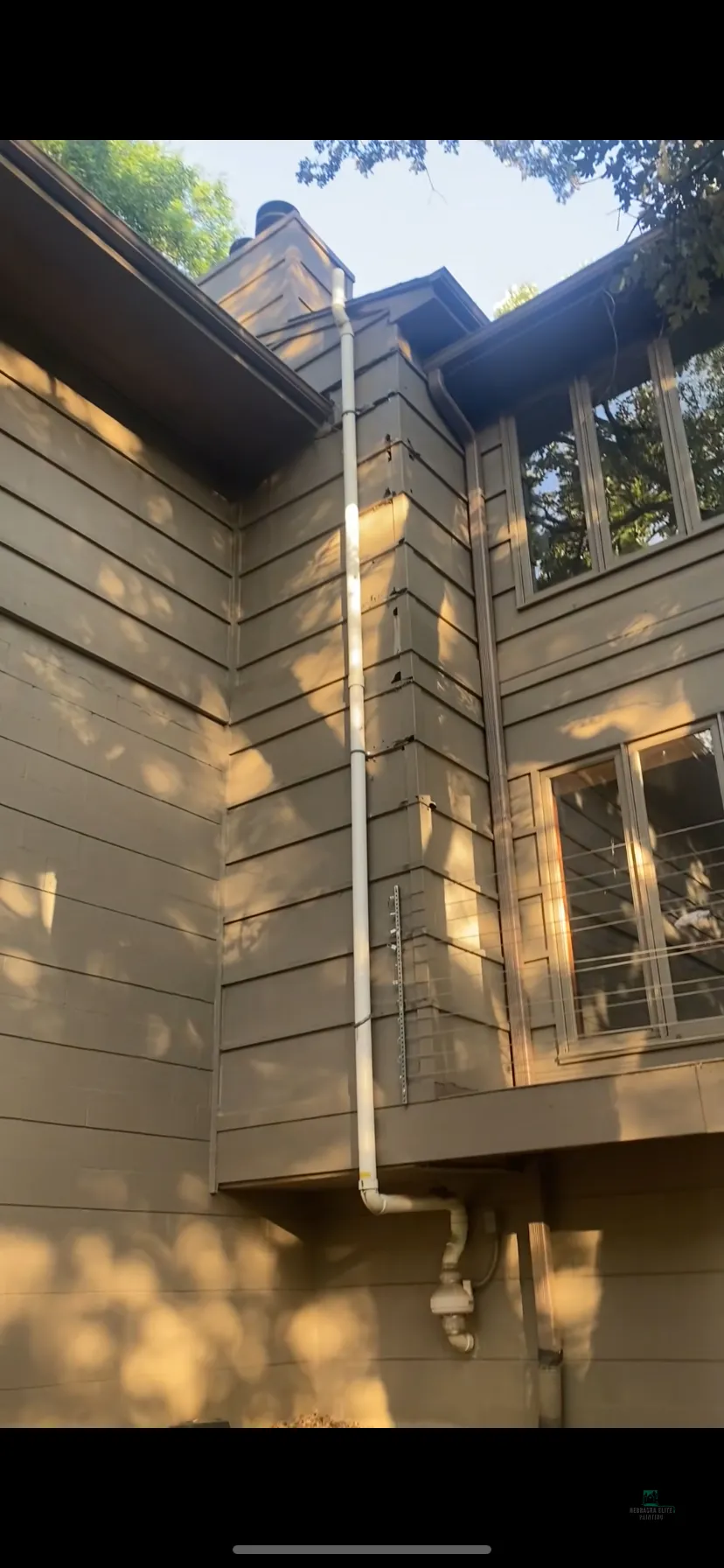 White downspout on brown wooden house exterior with sunlight filtering through trees.