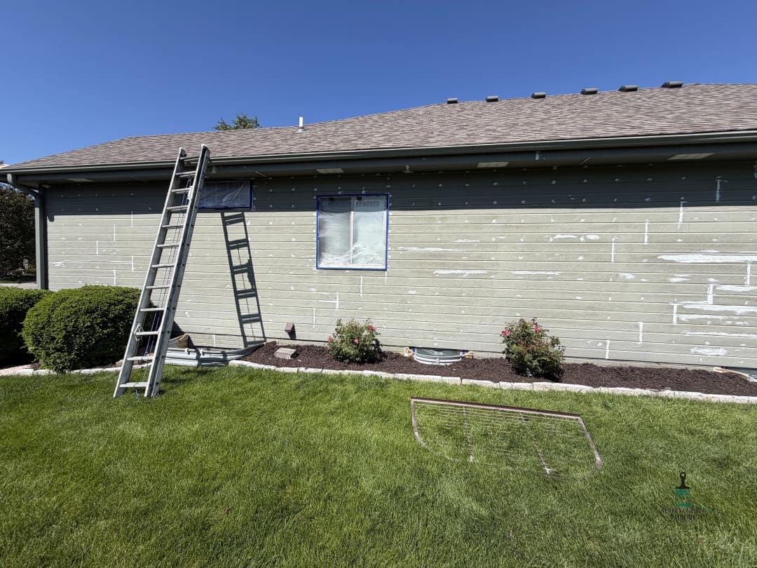 House exterior with partially painted siding, ladder leaning against wall, garden in foreground.