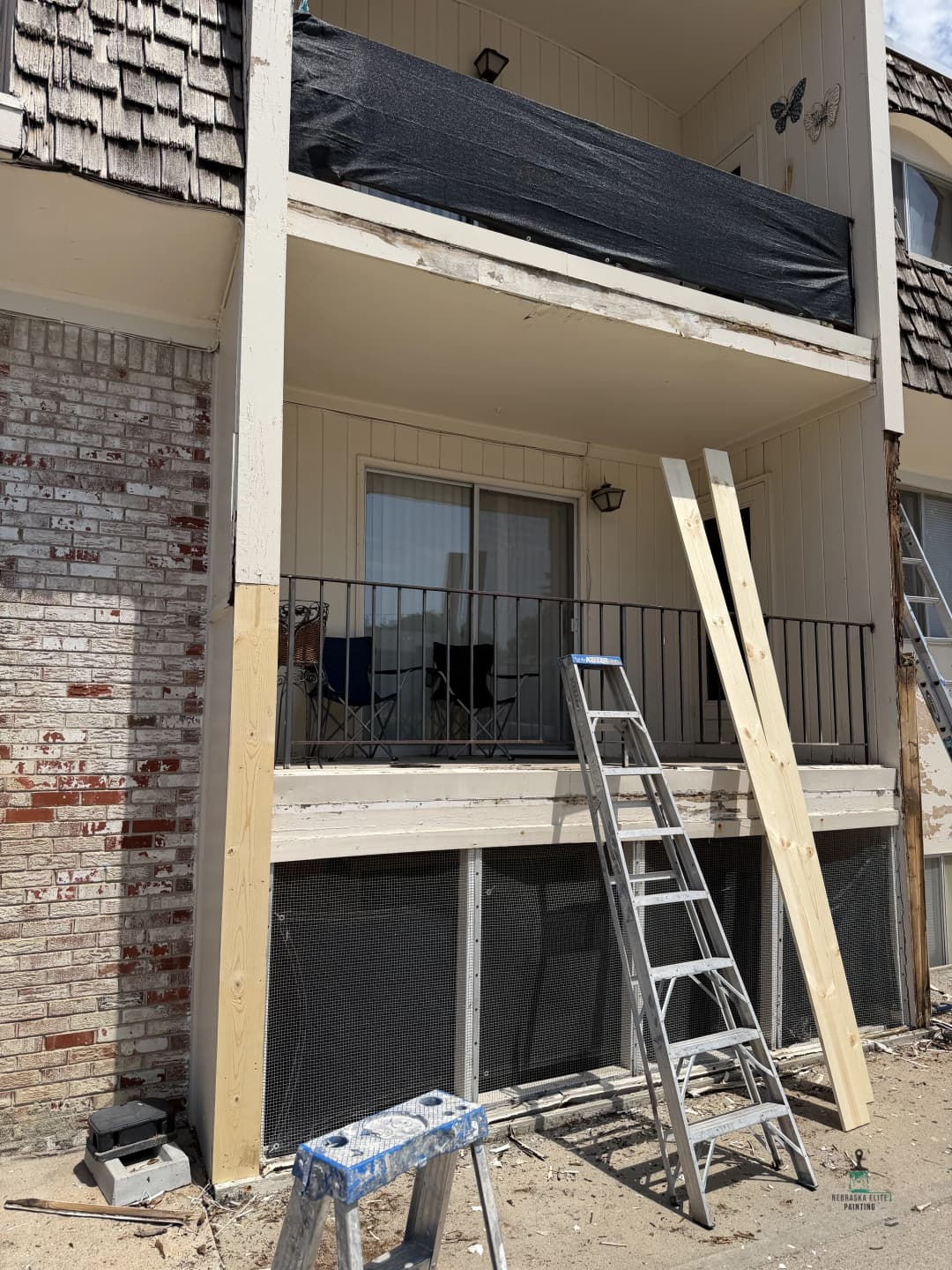 Balcony renovation with black tarp, ladder, and wooden supports at apartment building.