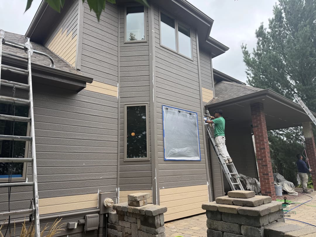 Man painting exterior of a house with ladder, gray siding, and preparation for window area.