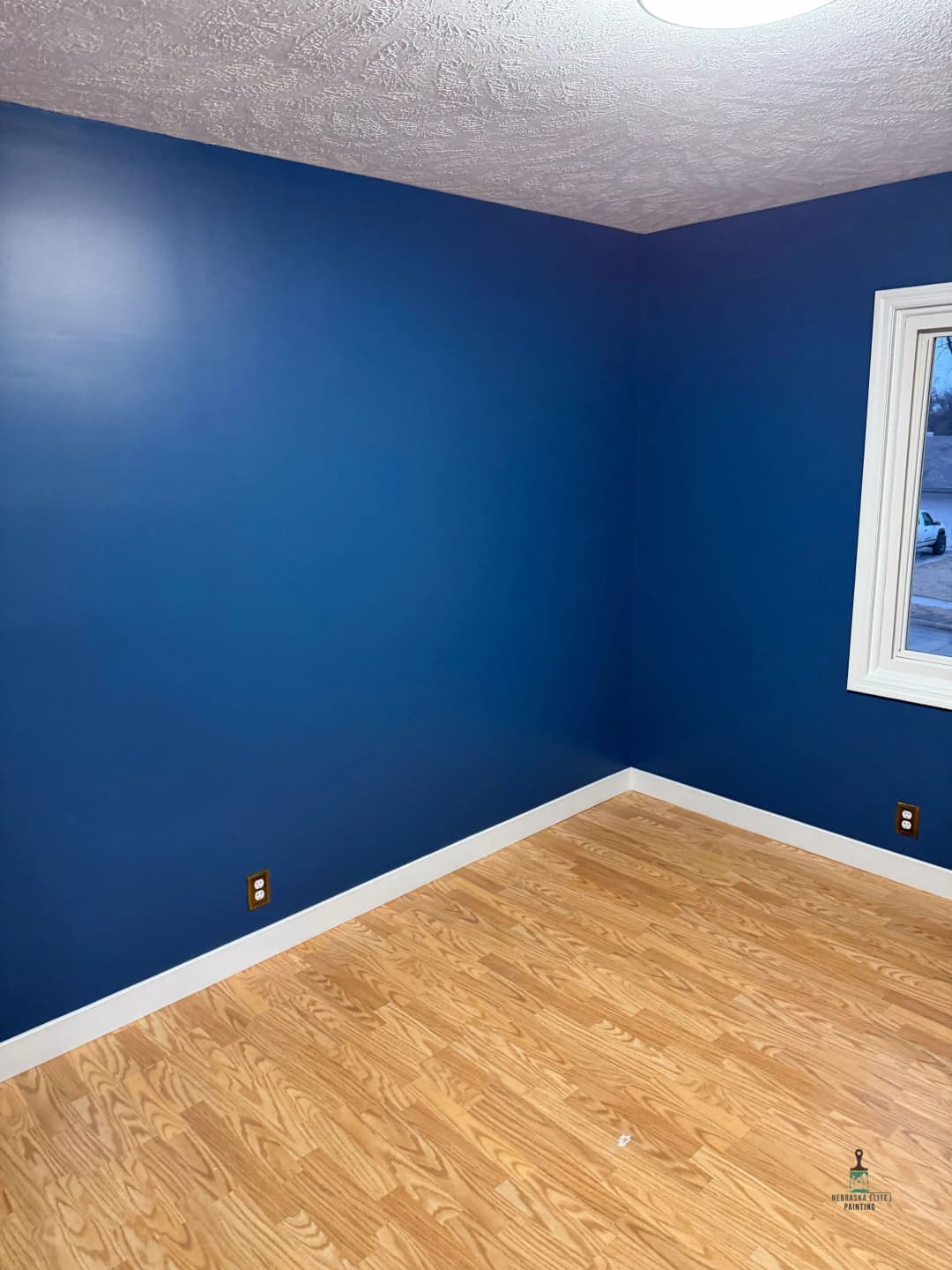 Interior of a room with blue walls and hardwood flooring, featuring a window and electrical outlets.