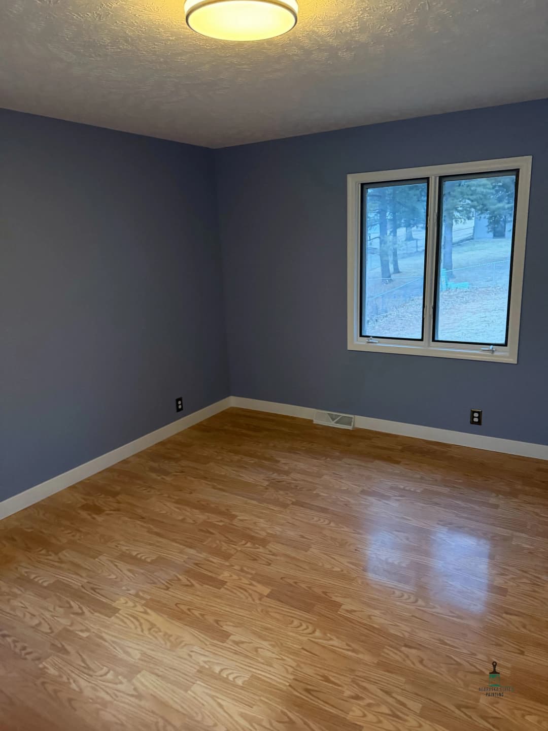 Empty room with light blue walls, hardwood floor, and large window overlooking trees.