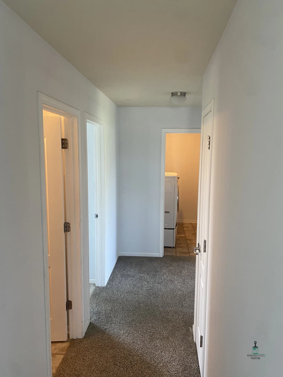 Bright hallway with beige carpet, leading to laundry room and two doors. Clean, simple design.