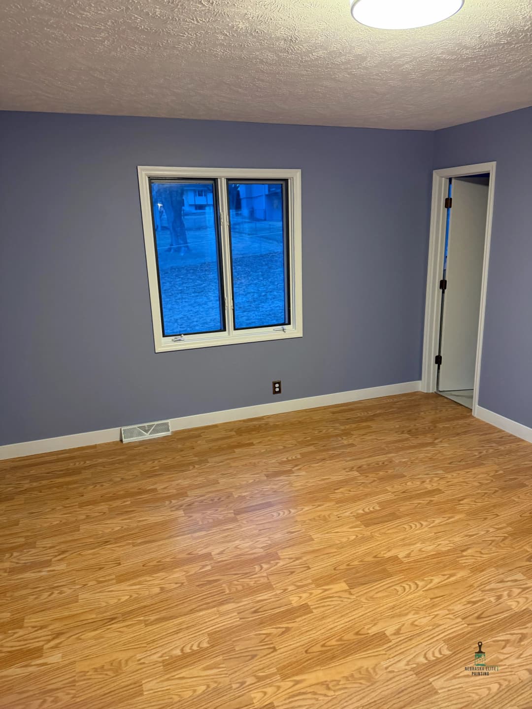 Empty room with light purple walls, hardwood floor, and large window overlooking outdoor view.