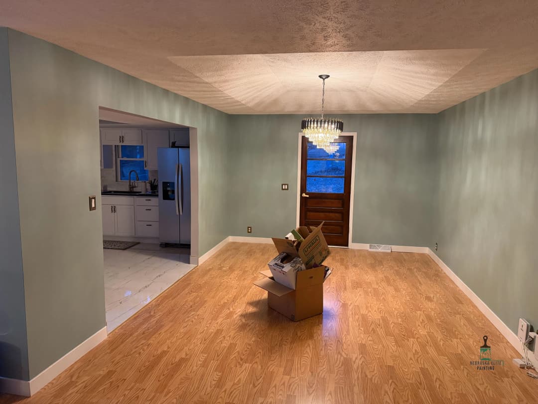 Empty living room with hardwood floors, light green walls, and a chandelier. Boxes in the center.