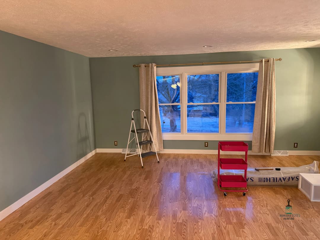Living room with fresh green walls, wooden floor, ladder, and red cart by large window.