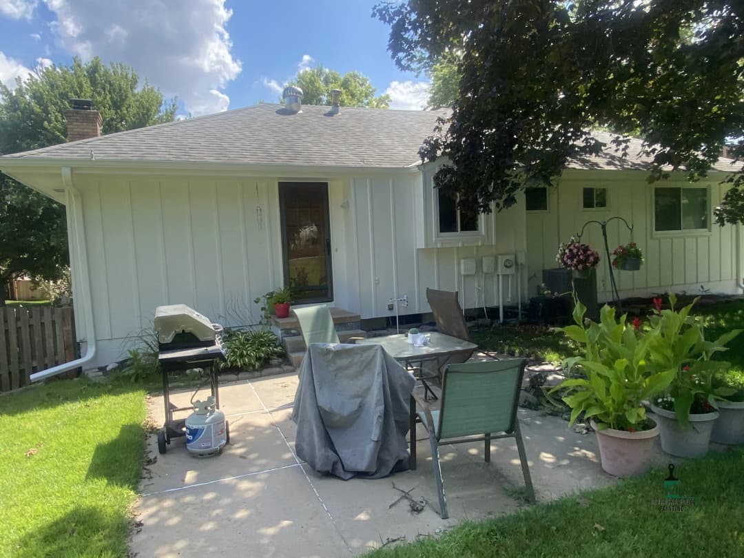 Backyard patio with grill, chairs, and flower pots at a white house on a sunny day.