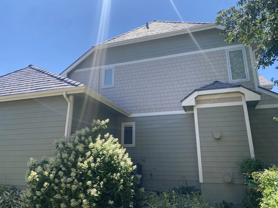 Two-story house with gray siding and shingles, surrounded by green shrubs and clear blue sky.