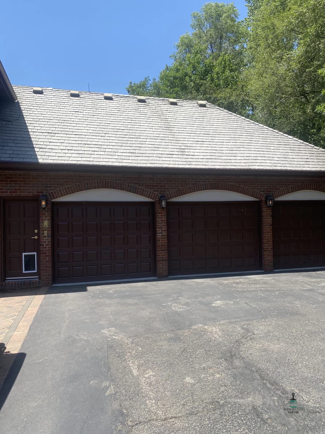 Three brown garage doors with brick accents under a clear blue sky and lush greenery.