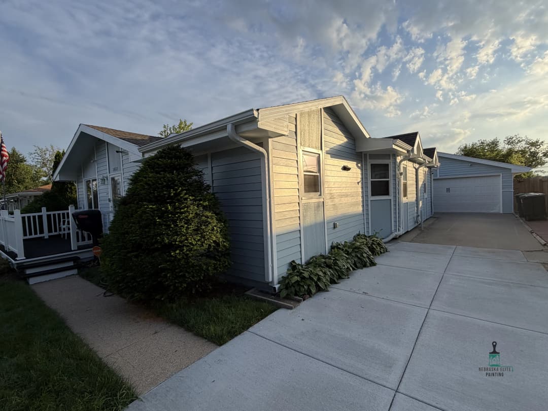 Single-story blue home with landscaped yard and driveway, under a cloudy sky.