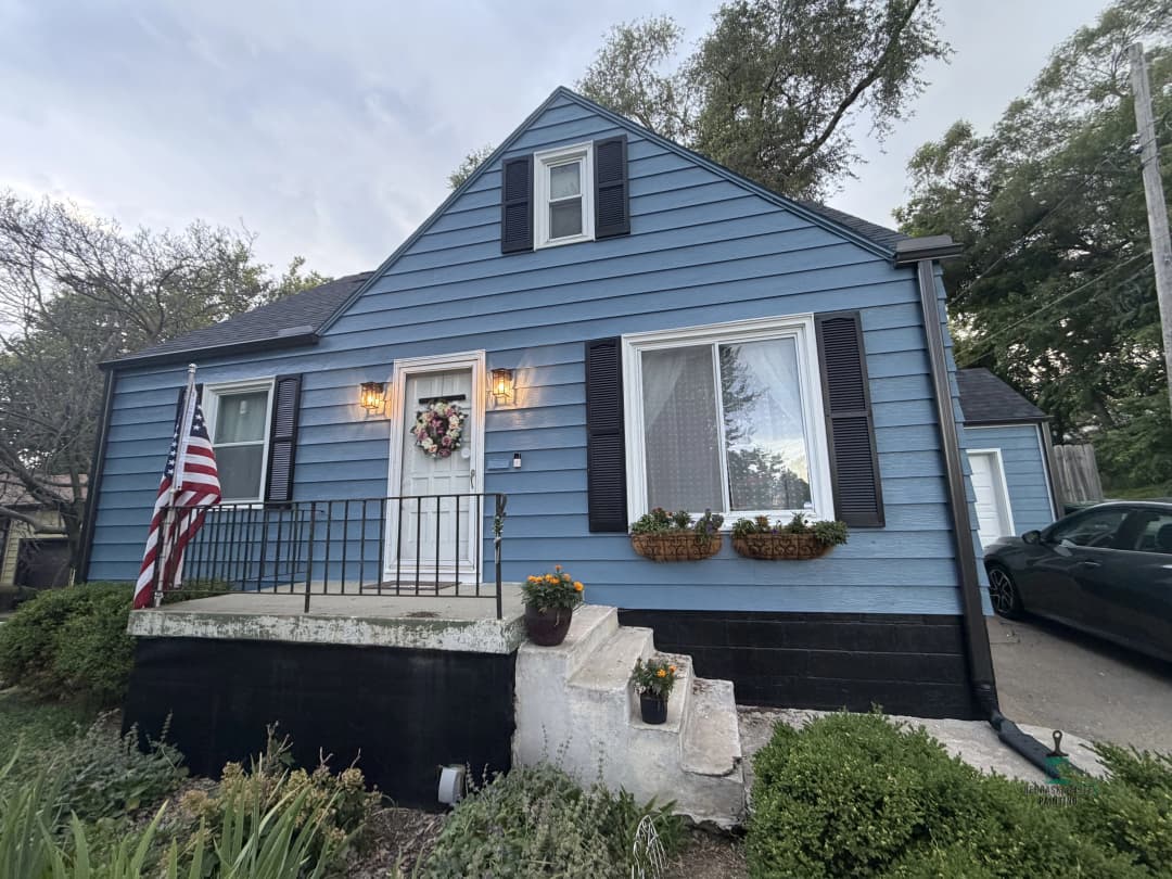Blue cape cod style house with front porch, American flag, and flower pots on windows.