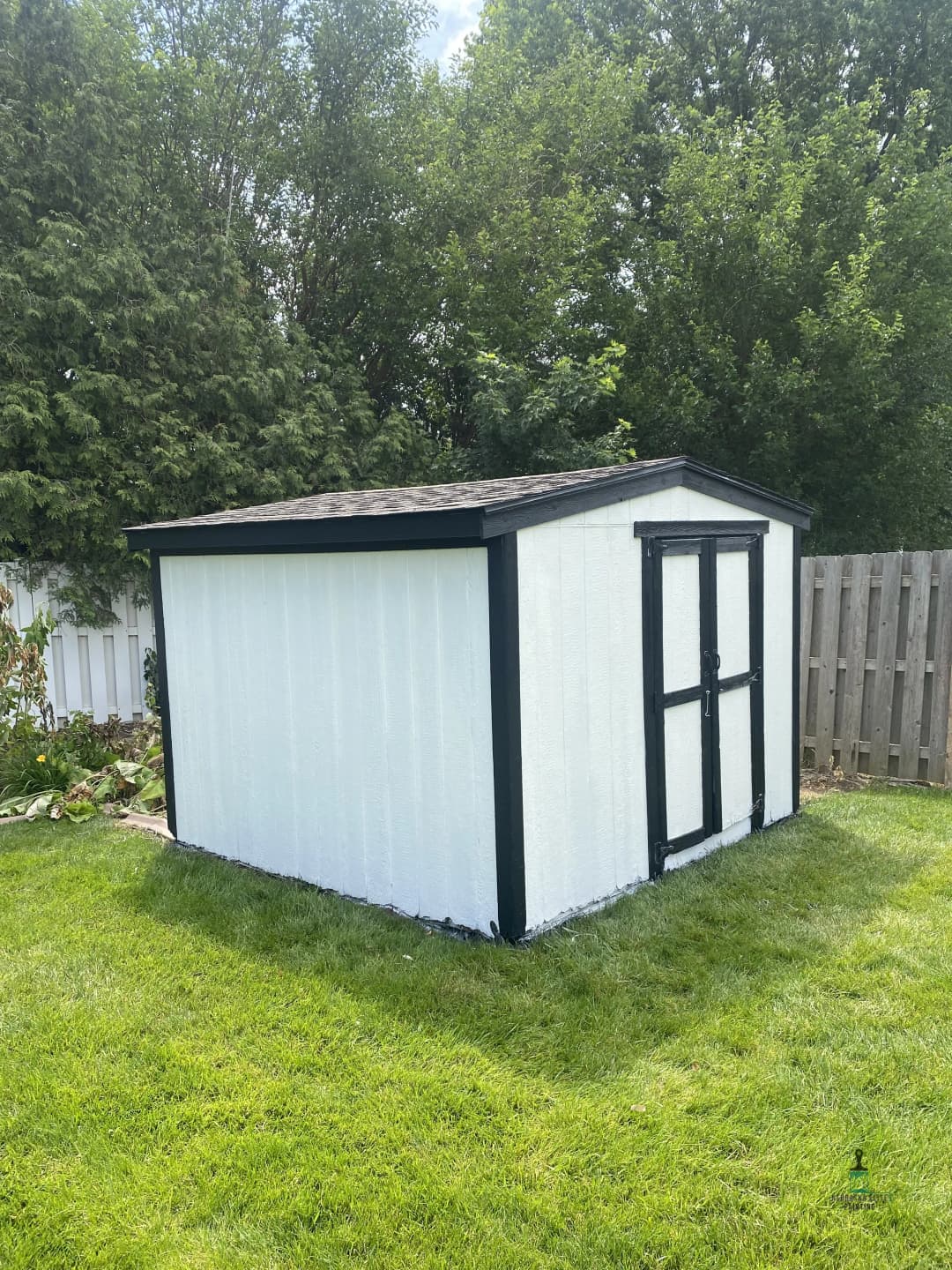 White outdoor shed with black trim in a green backyard surrounded by trees.