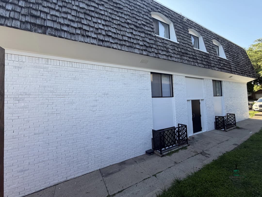 Exterior of a white brick building with a shingled roof and entrance, featuring black railings.