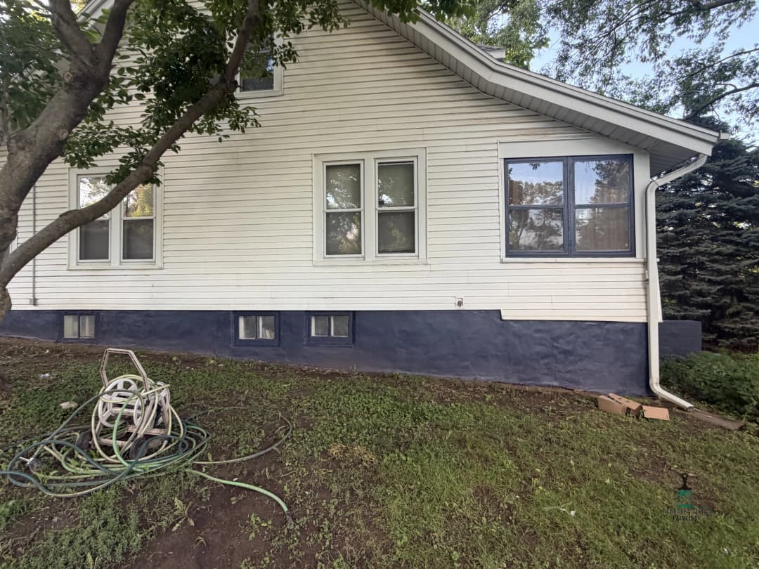 Exterior side view of a house with white siding and blue foundation, featuring lawn and garden hose.