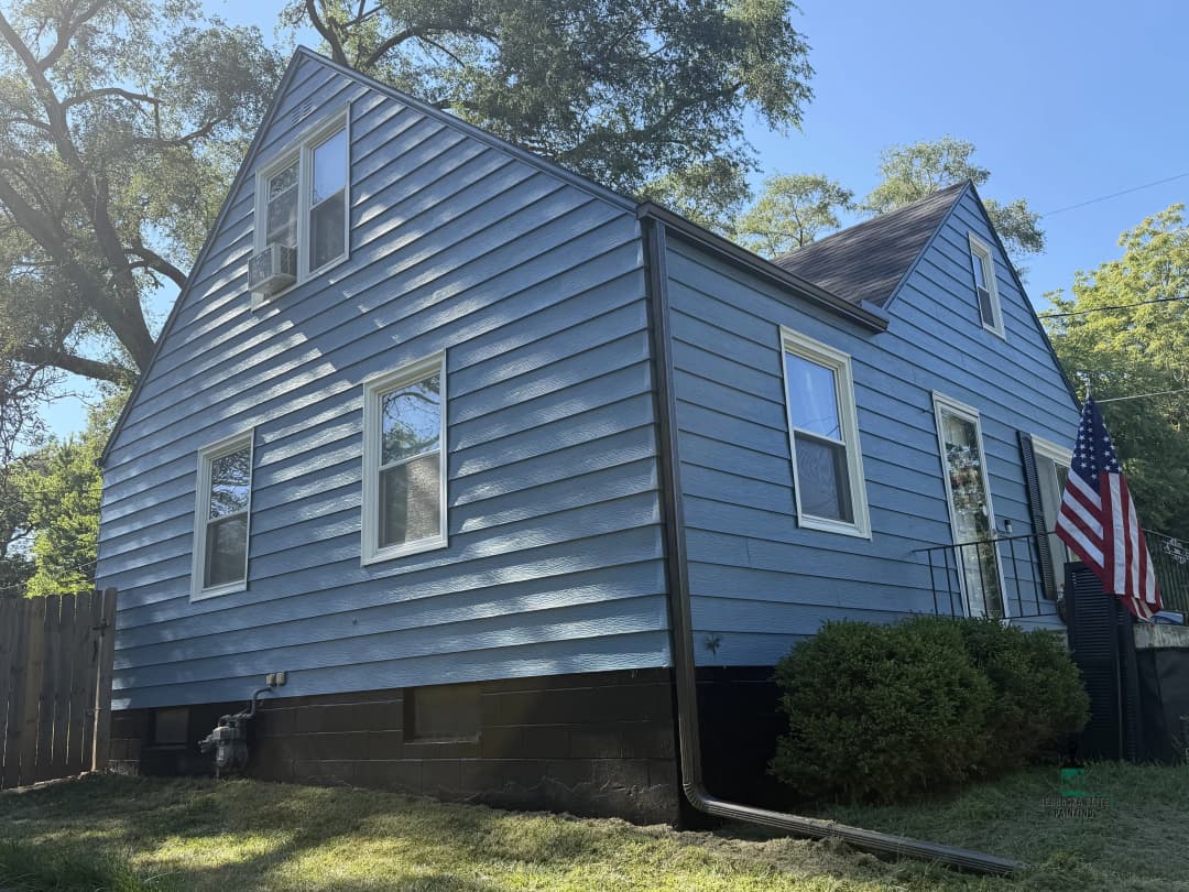 Blue house with American flag, side view, surrounded by greenery and clear sky.