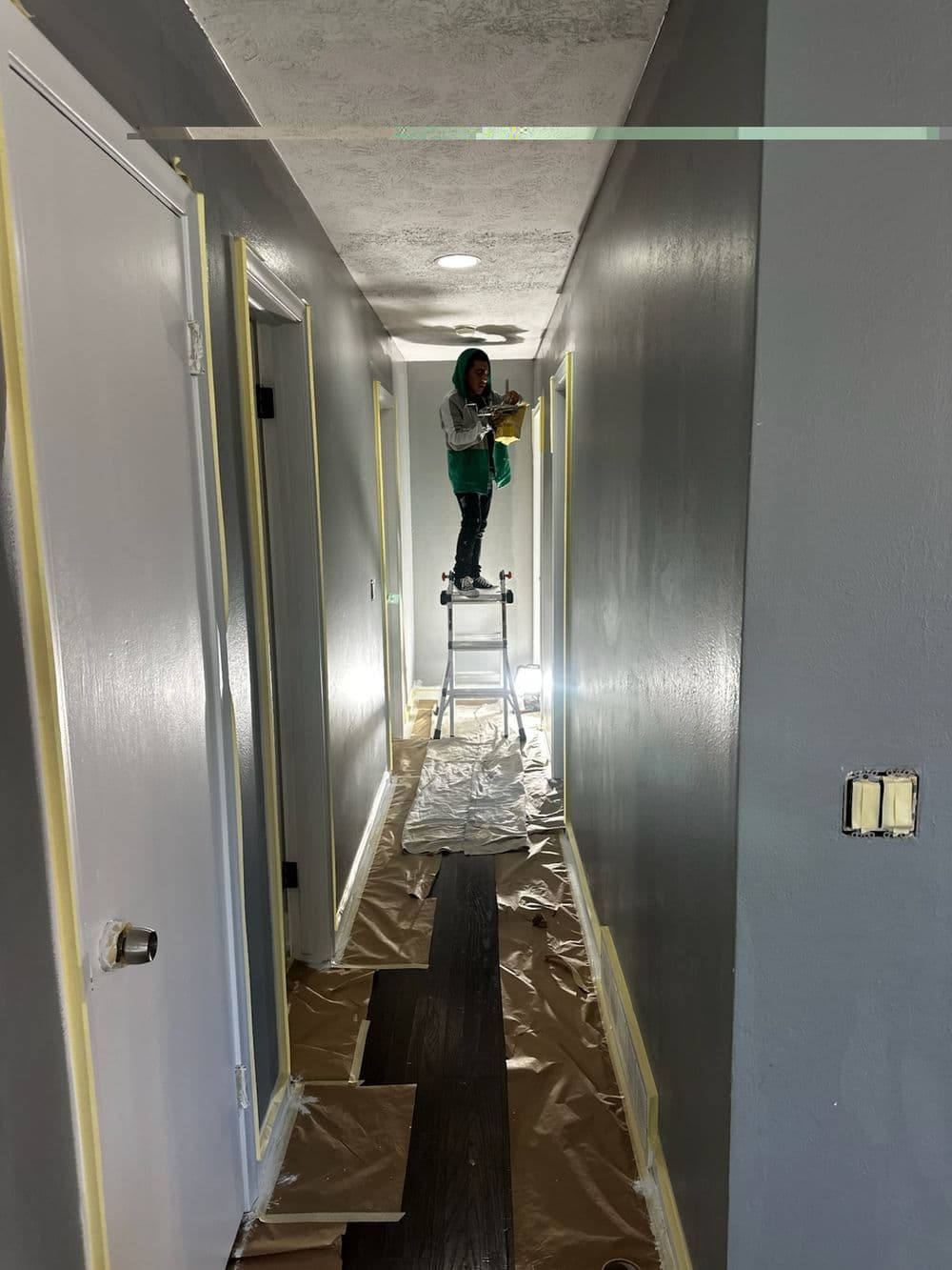 Person painting a hallway with yellow tape, standing on a ladder, protective covering on floor.