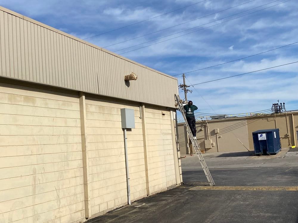 Worker on ladder performing maintenance on building exterior under clear blue sky.