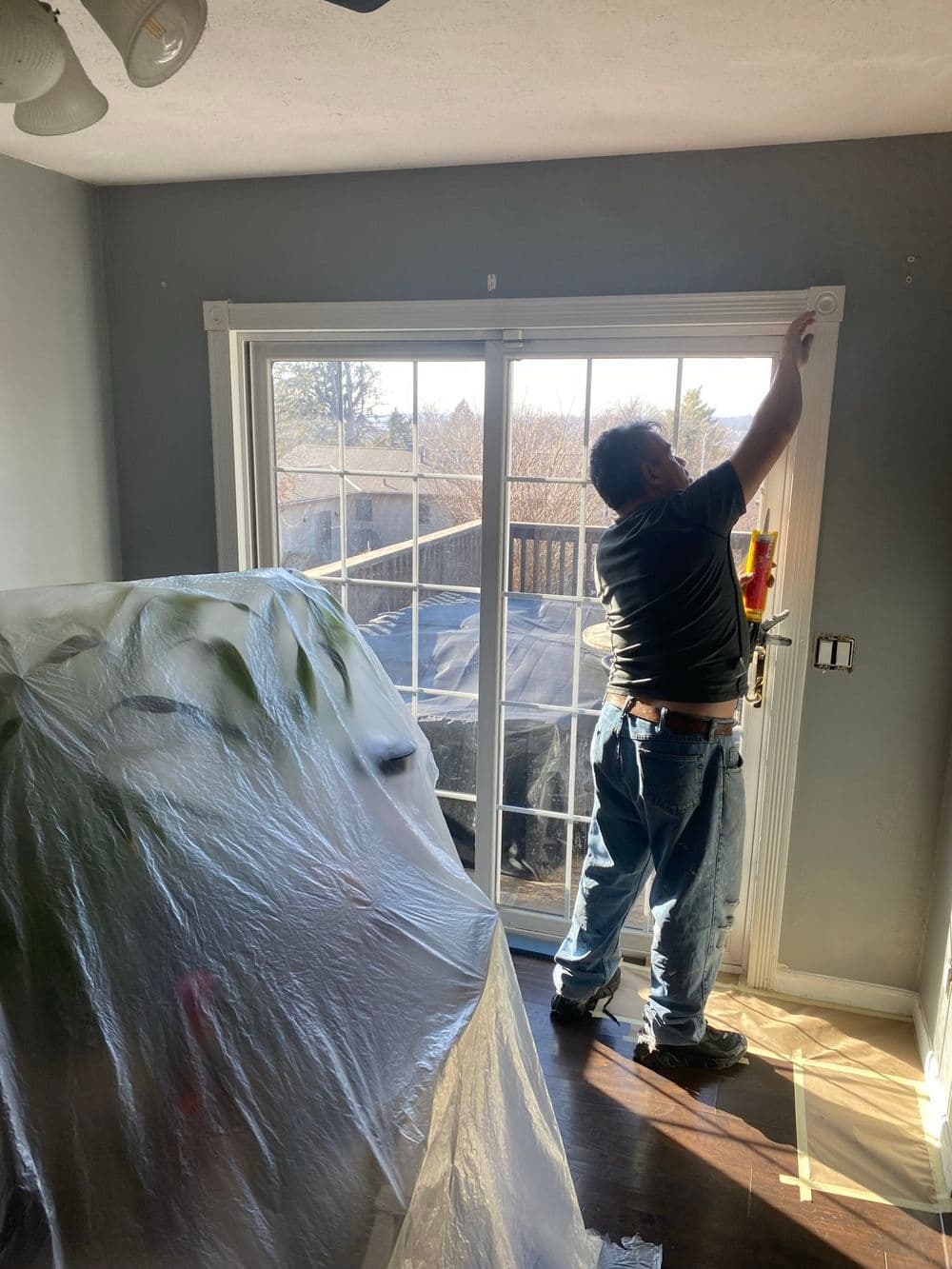 Man installing a door trim in a room with a plastic-covered furniture and natural light.