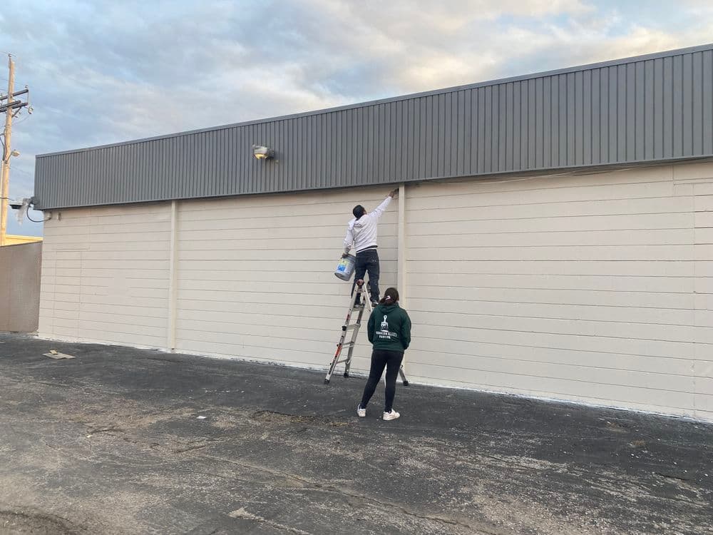 Person painting a building wall while another assists from the ground on a ladder setup.