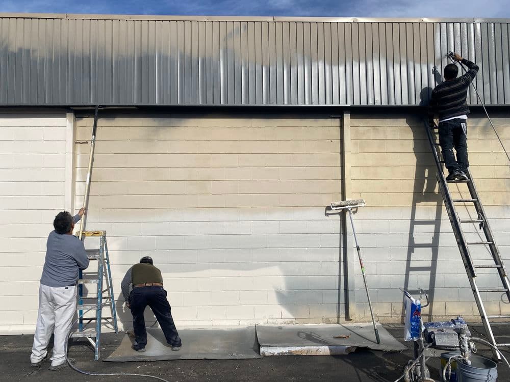 Three workers painting a building's exterior with ladders and scaffolding.