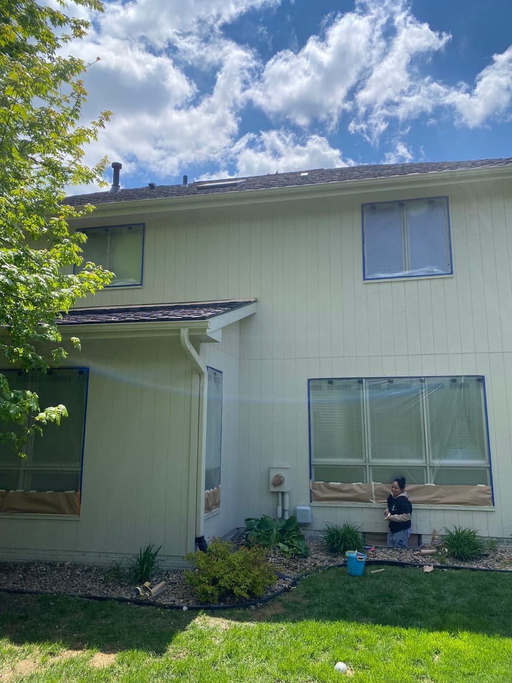 Person painting exterior wall of a house under a bright blue sky with fluffy clouds.