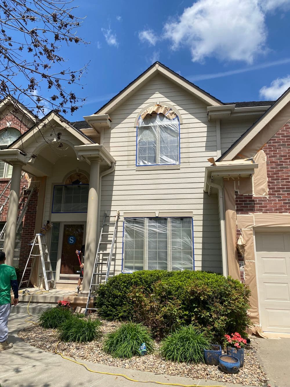 Home exterior renovation with ladders, masking, and landscaping on a sunny day.