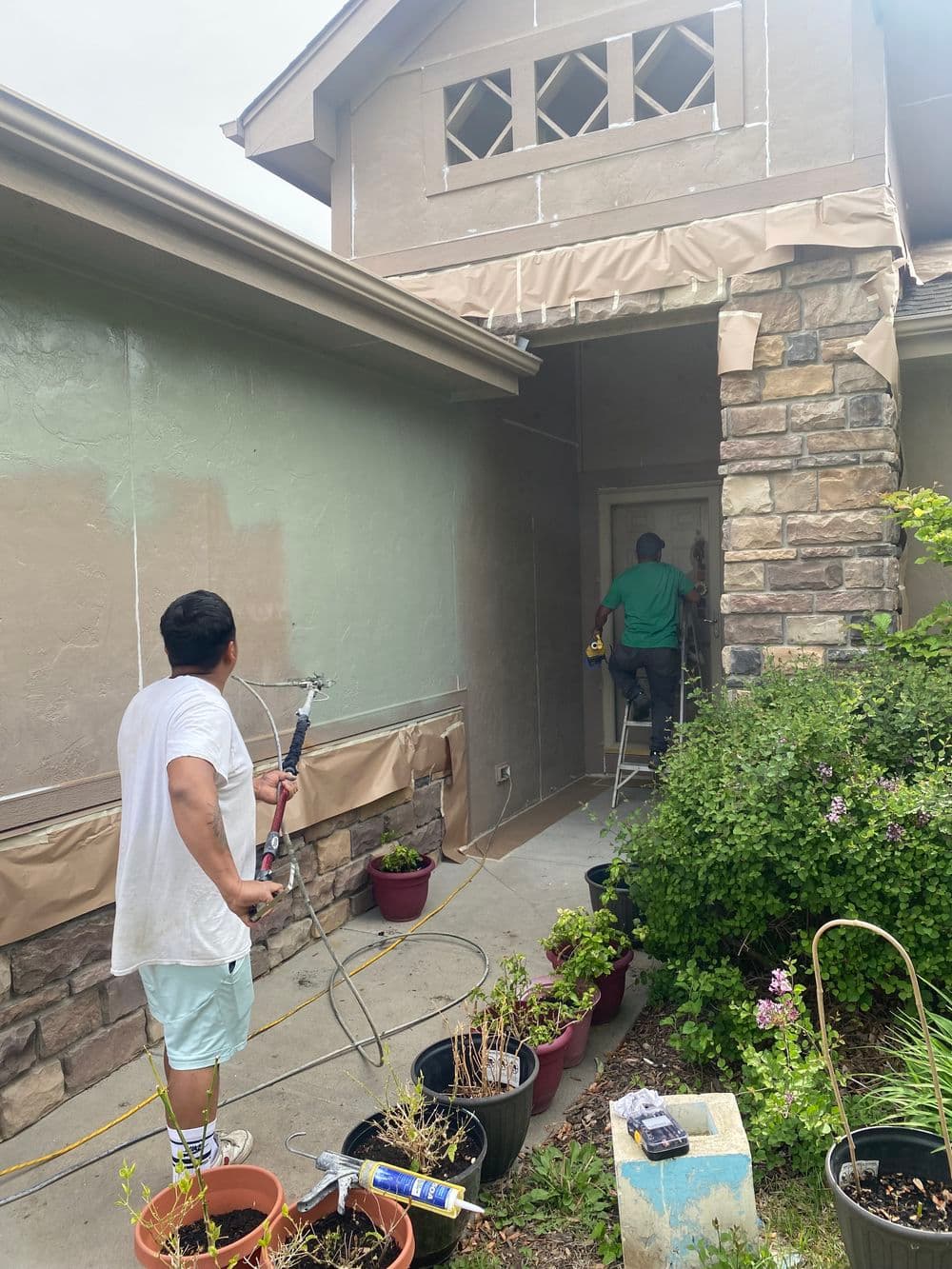 Home exterior painting: contractors painting house entry with sprayer and ladder, surrounded by plants.