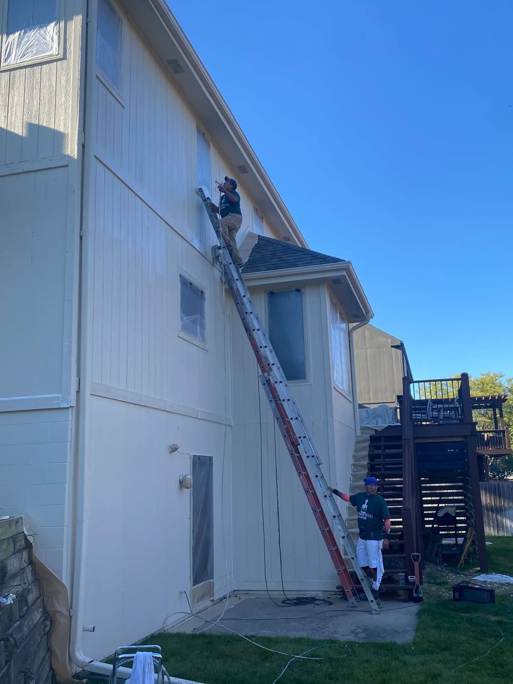 Two painters working on a house exterior, one on a ladder applying paint, clear skies above.