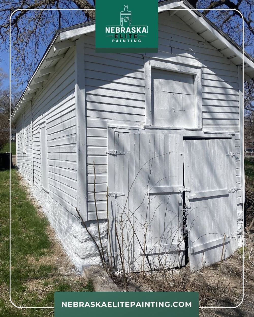 White-painted barn with wooden doors, surrounded by bare branches, Nebraska Elite Painting.