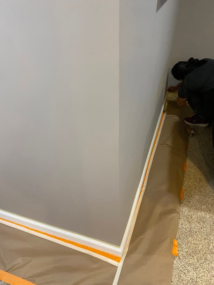 Person painting a wall corner with protective tape and drop cloth on the floor.