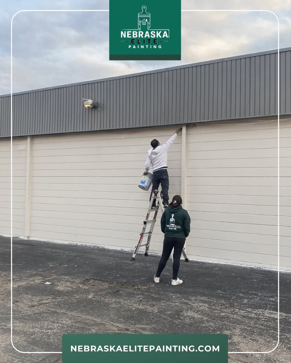 Worker painting a building exterior while another assists from the ground. Nebraska Elite Painting.